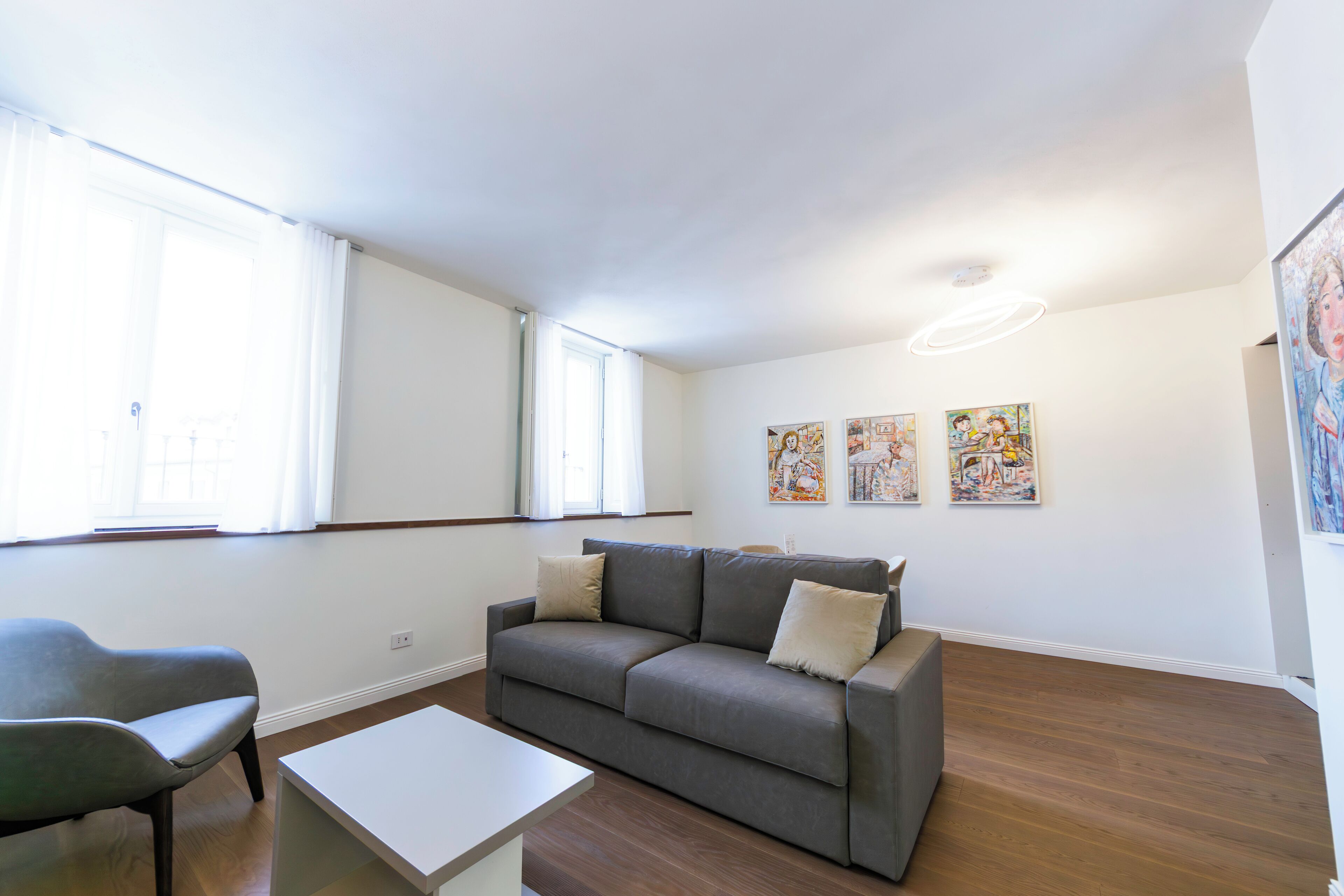 A modern living room with a grey sofa, a white coffee table, and a grey armchair. There are three paintings on the wall and a large window with white curtains.
