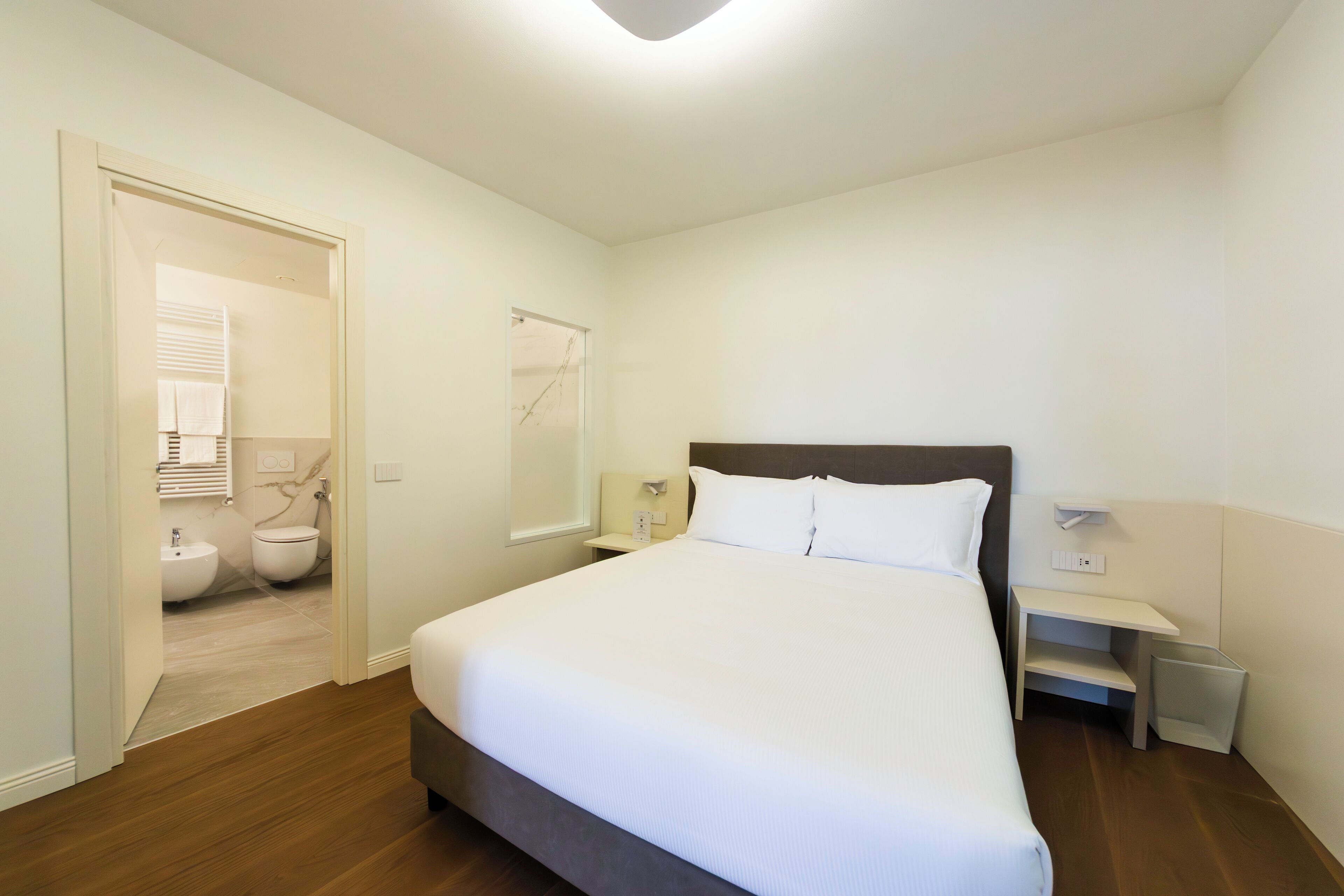 A bedroom with a large bed, a bedside table, a nightstand, a mirror, and a view of the bathroom.