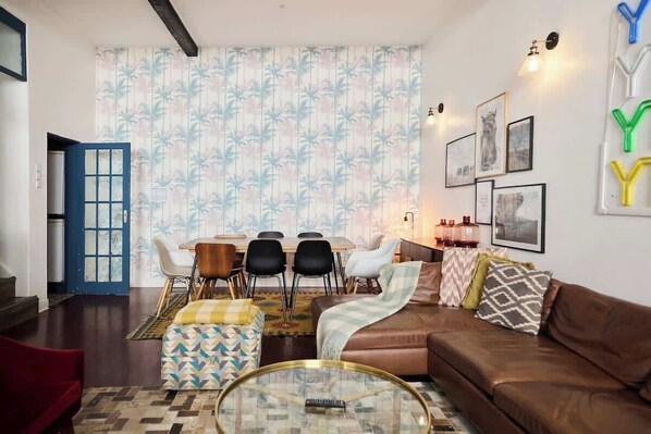 Un salon avec un canapé en cuir marron, une table basse en verre, un coin repas avec une table et des chaises, et un rideau à motifs.