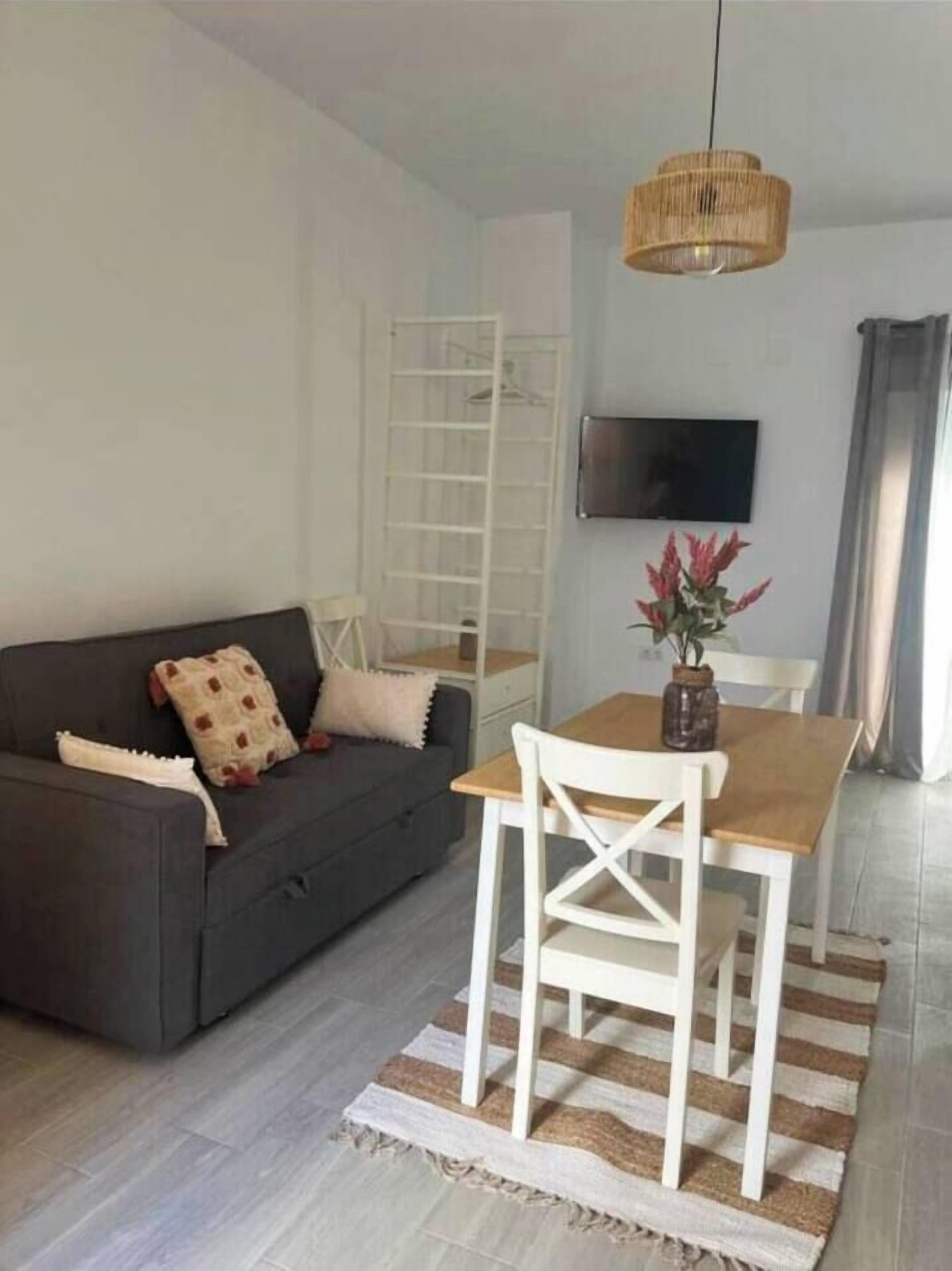 Una sala moderna con un sofá gris, una mesa de comedor de madera con sillas blancas, una lámpara colgante tejida y un televisor montado en la pared.