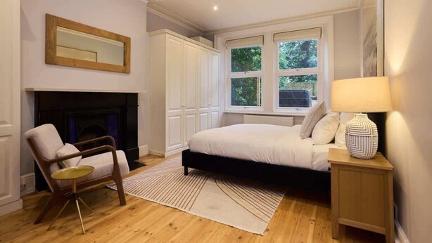 A bedroom with a bed, a chair, a fireplace, a nightstand, and a window with a view of greenery. - The West Kensington Hideout (Greater London)