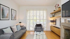Un salon comprenant un canapé, un fauteuil, une table basse, une cheminée et une grande fenêtre avec des stores.