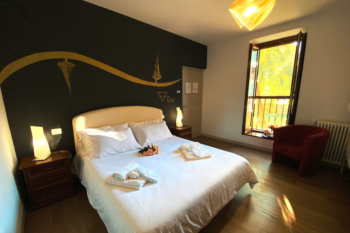 Un dormitorio con una cama grande, dos mesitas de noche con lámparas, un sillón rojo y una ventana con vistas a la vegetación.