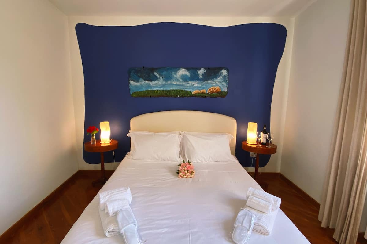 Un cuarto de hotel con una cama grande, dos mesitas de noche con velas, un cuadro en la pared y un ramo de flores.