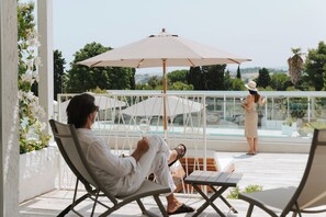 A man relaxing on a lounge chair under a large umbrella on a balcony with a view of a pool and greenery. - Nu Aria Taunus Relais  (Numana)