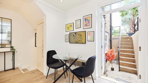 Un espace repas moderne avec une table en verre et des chaises noires, un escalier menant à un balcon et des œuvres d’art accrochées aux murs.