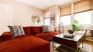 A living room with a sectional sofa, a coffee table, a mirror, and a window with blinds. - The Waterlow Park Wonder - 2BDR with Study (Greater London)
