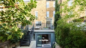 Un apartamento moderno con entrada por puerta de cristal, un pequeño espacio de asientos al aire libre y una escalera que conduce a un piso superior.