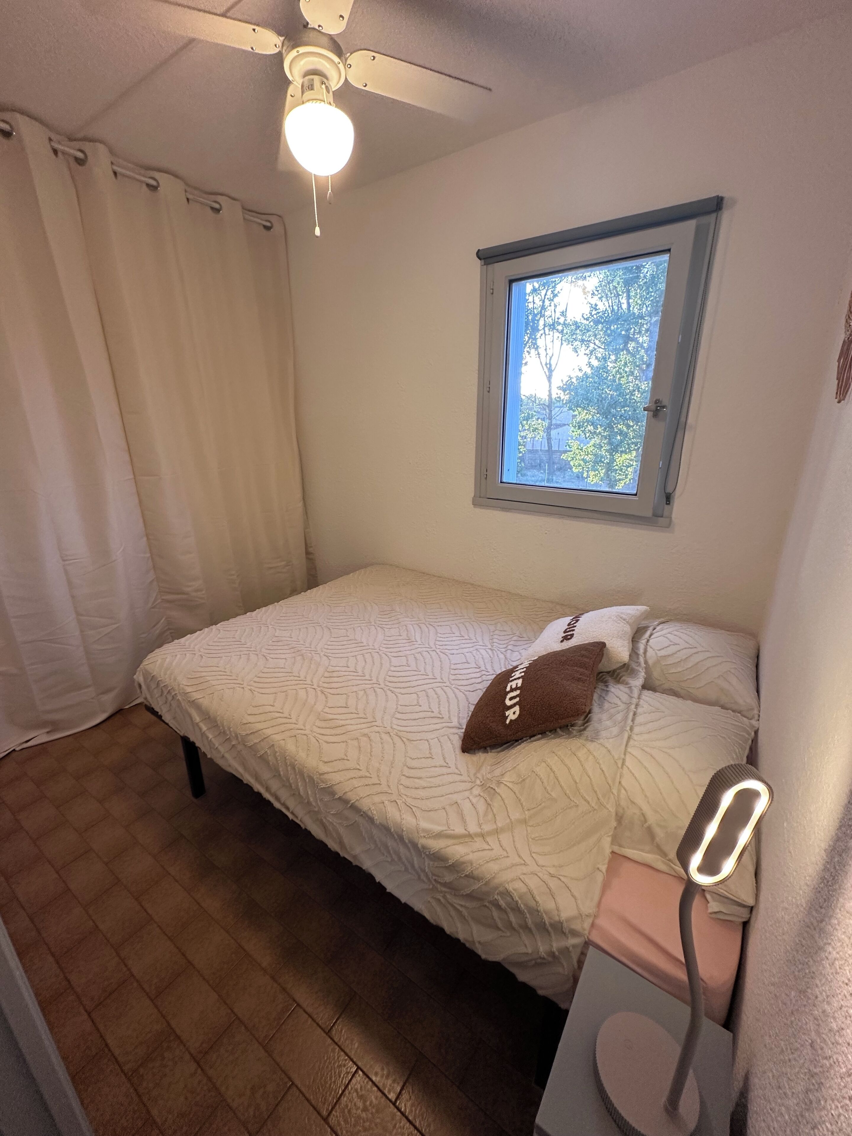 Une chambre à coucher avec un lit, une fenêtre, un ventilateur de plafond et une table de chevet.