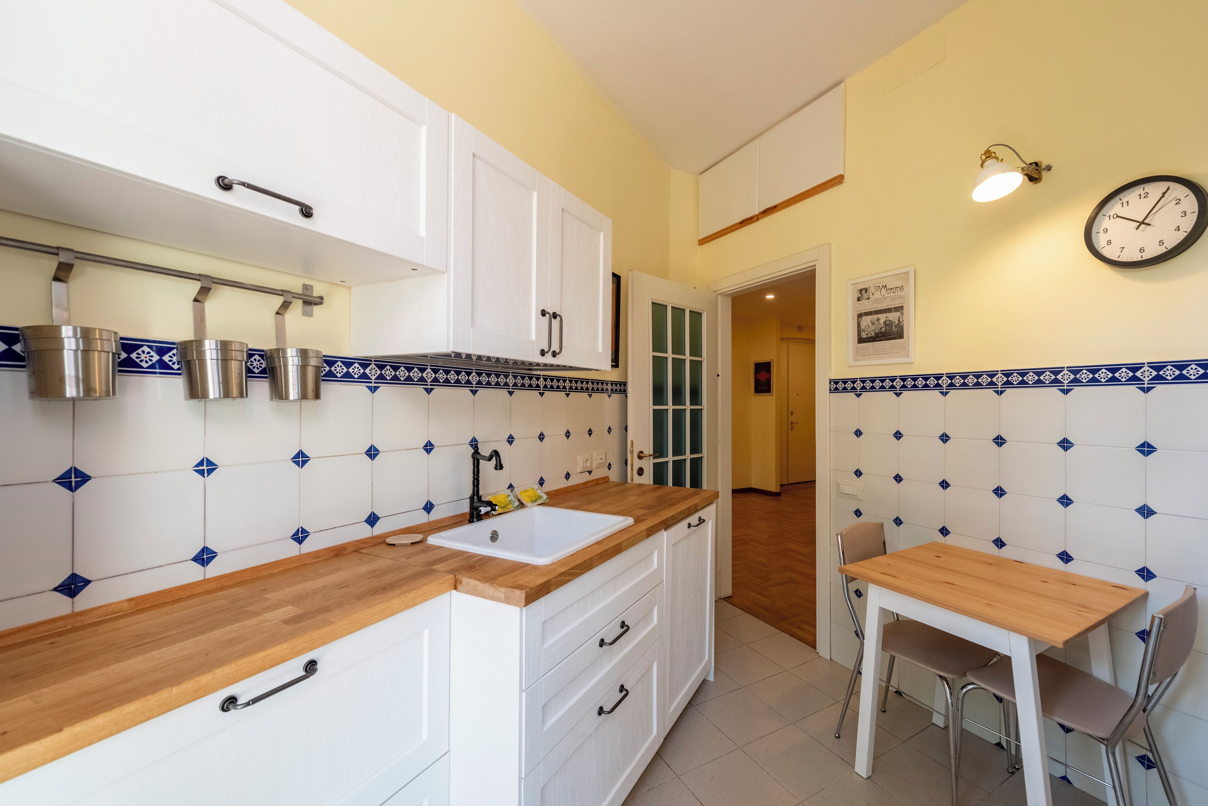 Une cuisine avec des meubles blancs, un plan de travail en bois et un évier. On y trouve une table à manger avec des chaises et une horloge murale.