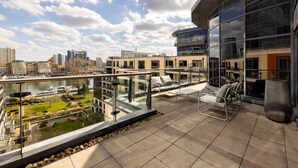 A modern building with a glass balcony overlooking a river, with seating and a view of the cityscape.
