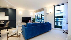 A modern living room with a blue sofa, a glass coffee table, a TV, and a large window with a view. - The Melbray Escape - Trendy 2BDR Apartment (Greater London)