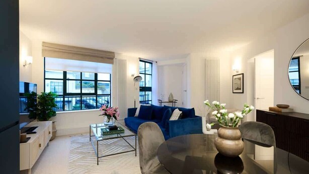 A modern living room with a blue sofa, a glass coffee table, and a dining table with chairs. There is a large window providing natural light and a view of the city. - The Melbray Escape - Trendy 2BDR Apartment (Greater London)
