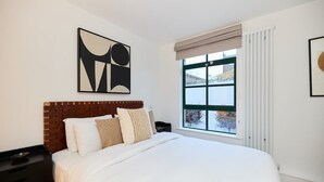 A bedroom with a large bed, a nightstand, a window with a view, and abstract wall art. - The Melbray Escape - Trendy 2BDR Apartment (Greater London)