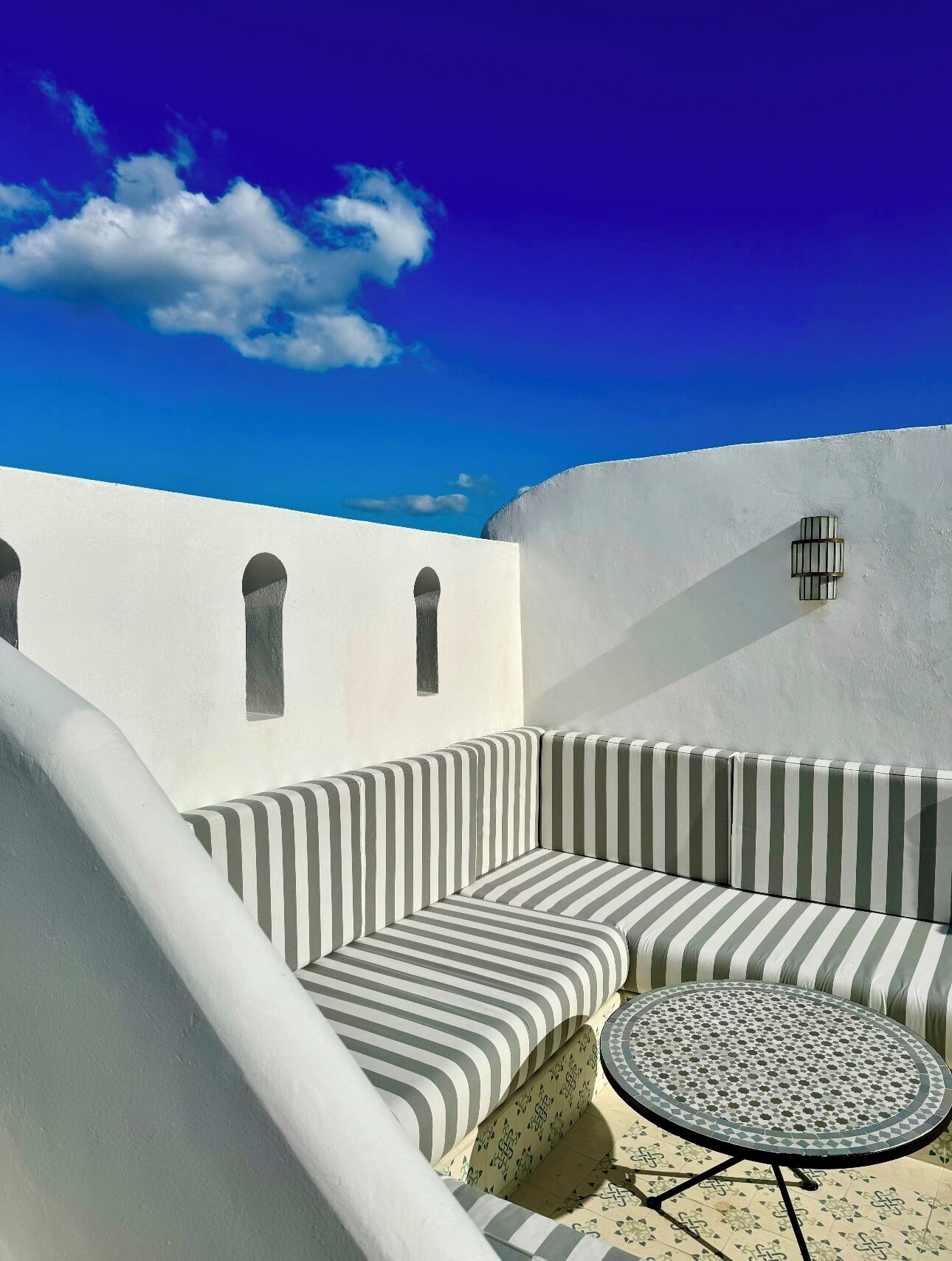 Un espace extérieur lumineux, aménagé avec un canapé à rayures et une table ronde, adossé à un mur blanc orné de fenêtres en arc de cercle et d’une petite fenêtre à barreaux.