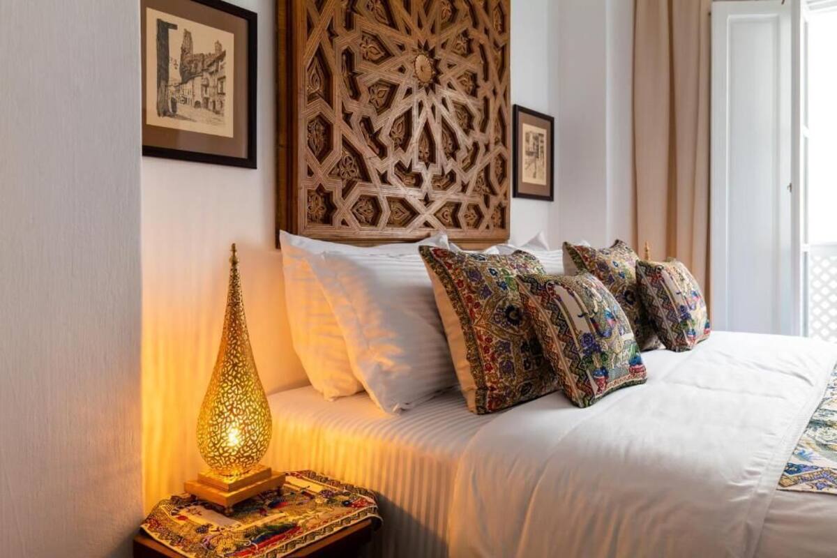 Ein ordentlich bezogenes Bett mit weißen Leinen und gemusterten Kissen, eine dekorative Wandverkleidung und eine Wandlampe.