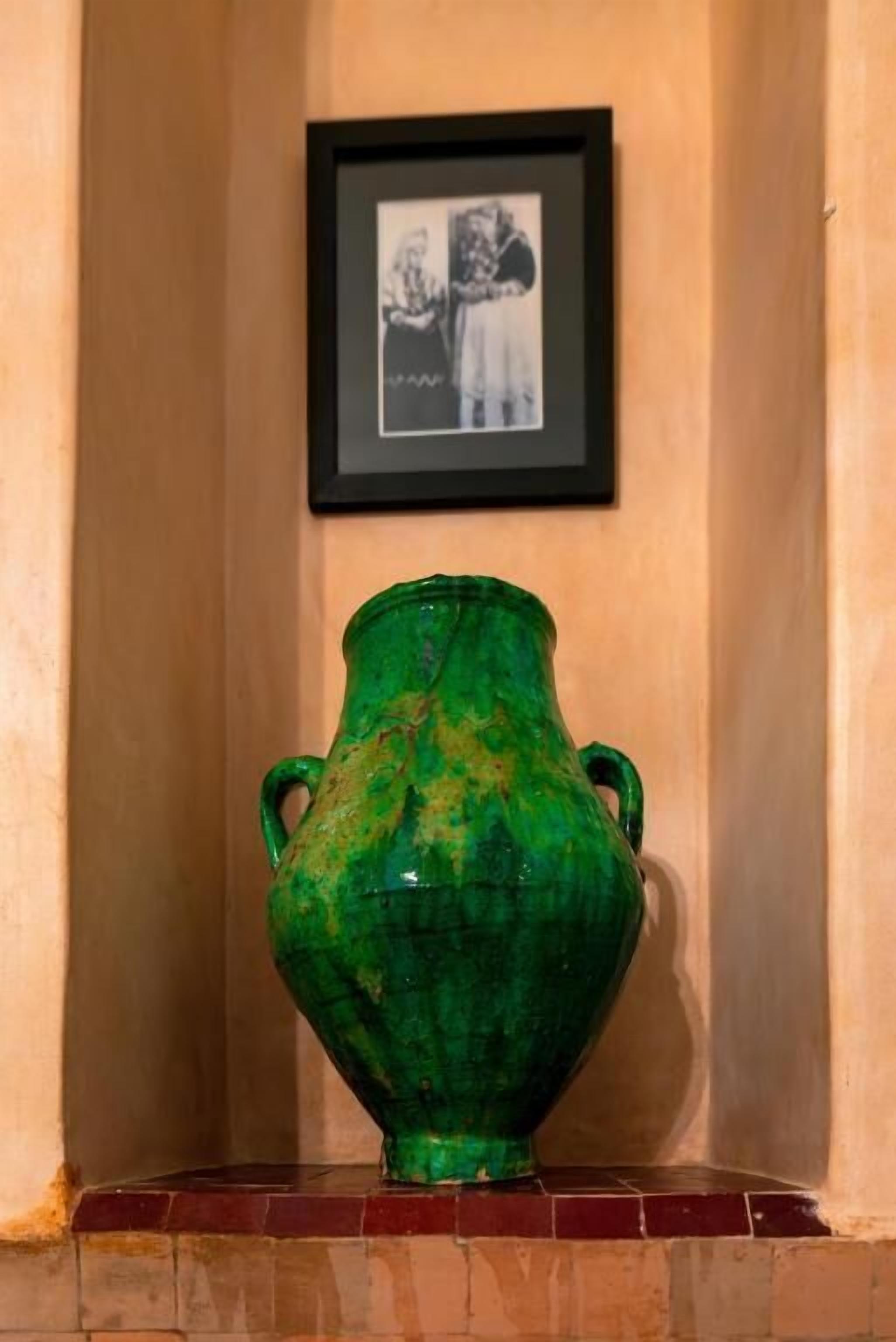 A green ceramic vase with two handles on a tiled ledge against a wall with a framed black-and-white photograph above it.
