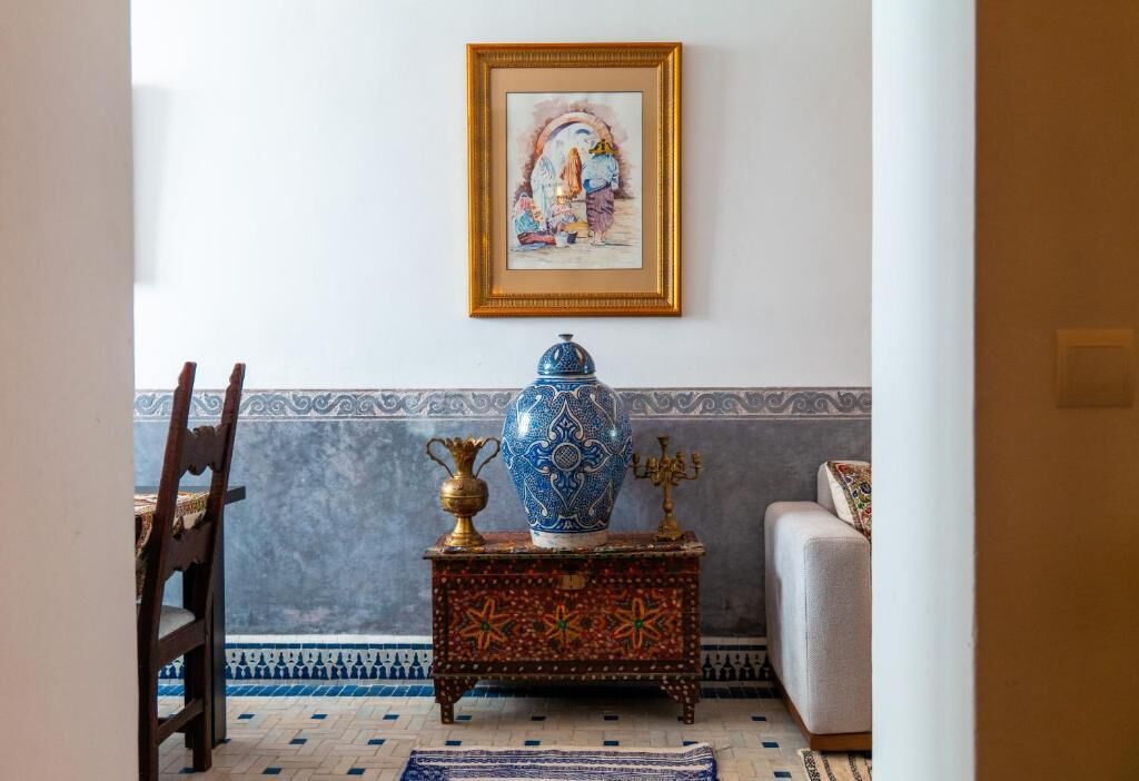 Una habitación con un alfombrado estampado, un jarrón azul y dorado, una mesita y un cuadro en la pared.