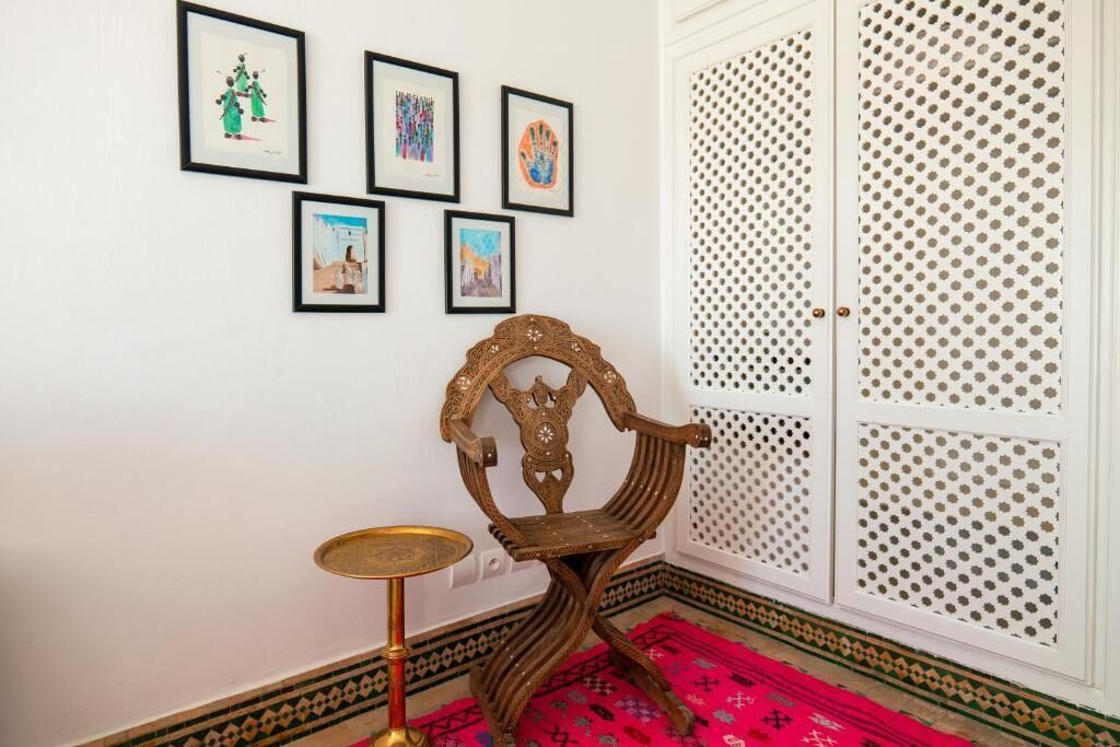 A room with a traditional wooden chair, a small round table, and a red patterned rug. There are four framed artworks on the wall.