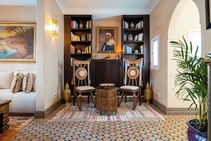 A traditional interior with a patterned floor, a central table with chairs, bookshelves, and a painting on the wall.