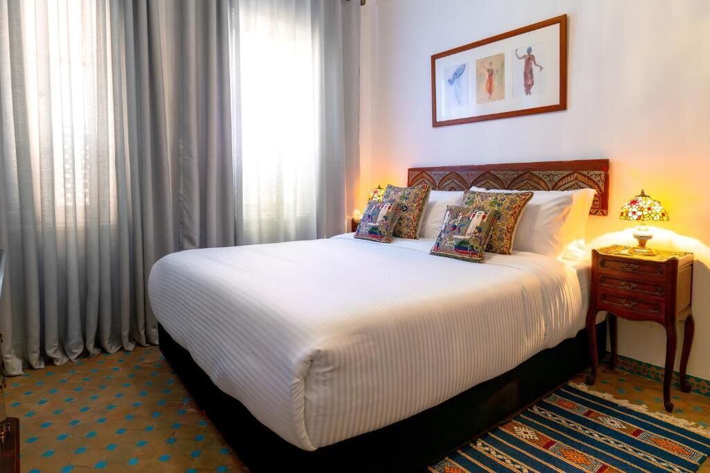Una habitación de hotel con una cama grande, una mesita de noche con lámpara y un ventanal con cortinas.