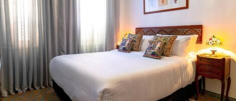Una habitación de hotel con una cama grande, una mesita de noche con lámpara y un ventanal con cortinas.