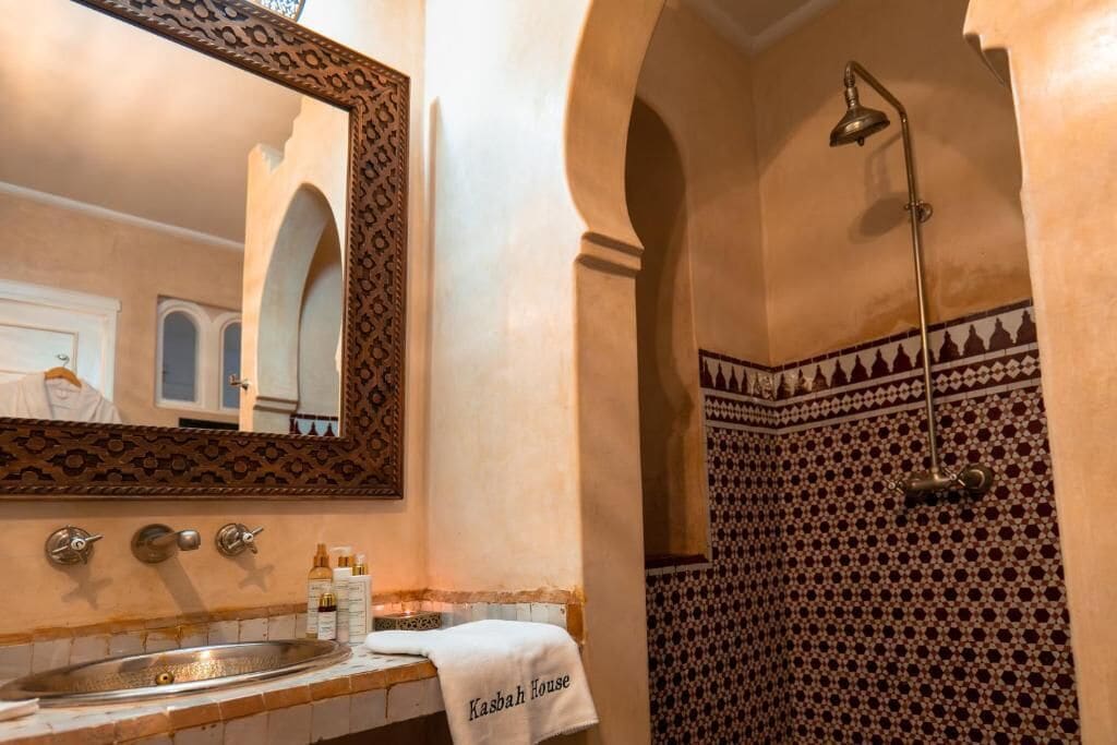 Casa de banho com um espelho grande, lavatório com artigos de higiene e zona de duche com azulejos estampados.