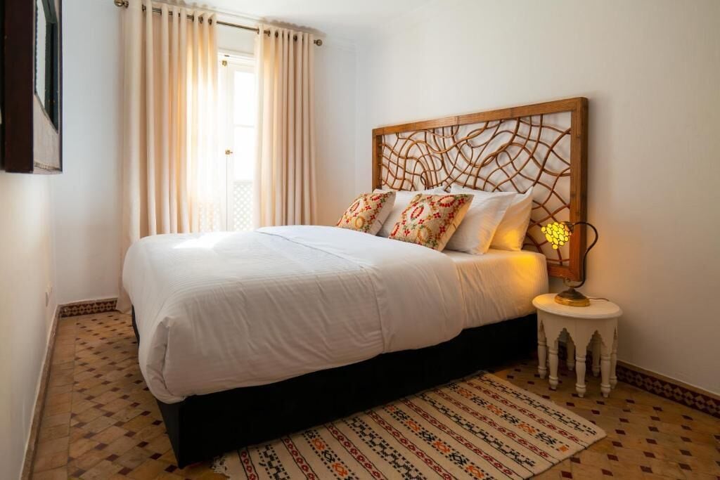 A bedroom with a bed, a patterned rug, a bedside lamp, and a window with curtains.