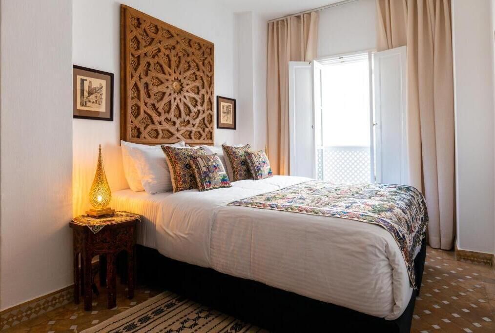 A bedroom with a large bed, a decorative wall panel, a nightstand with a lamp, and a window with curtains.