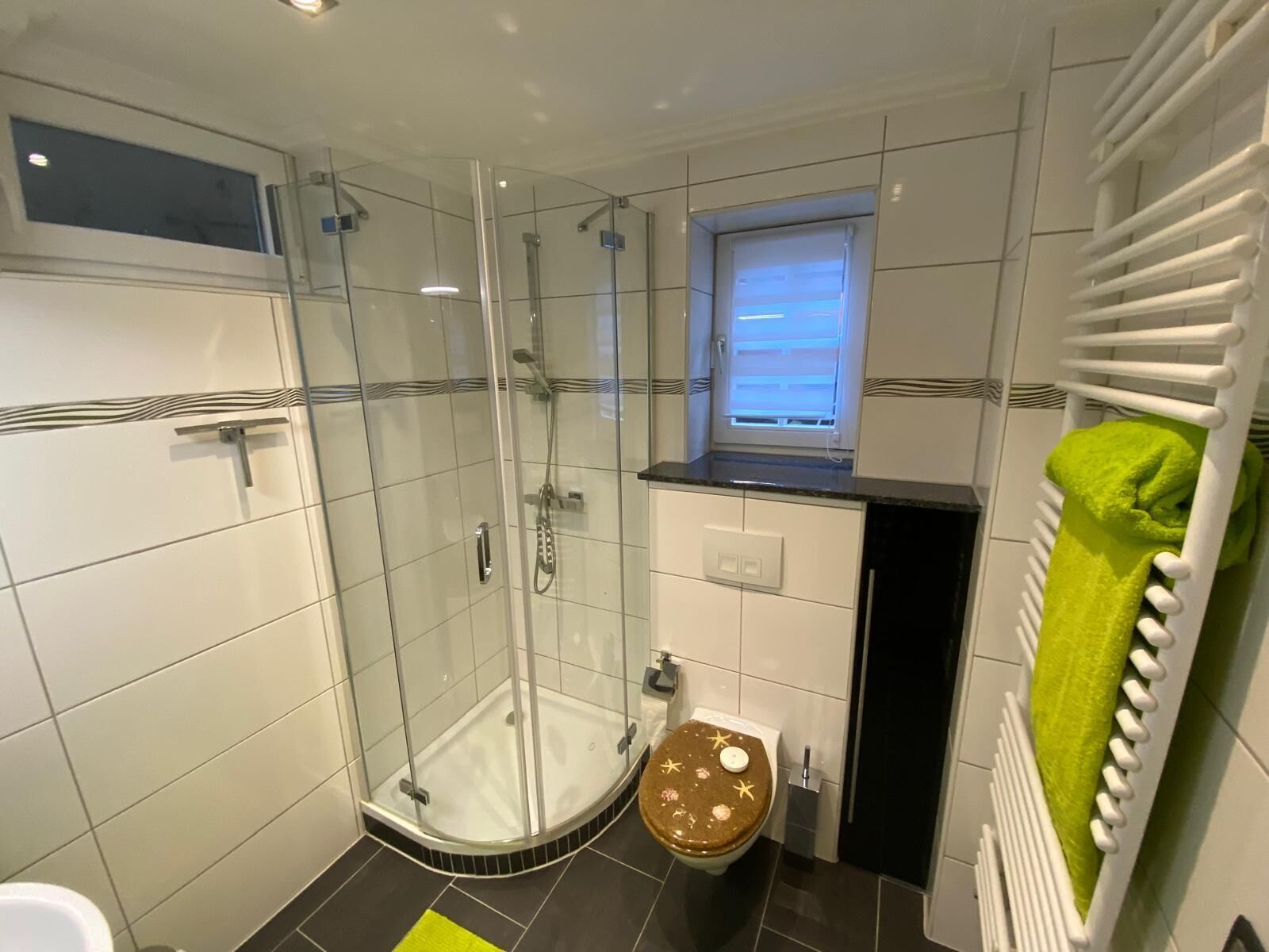 A modern bathroom with a glass-enclosed shower, a toilet with a decorative lid, and a towel rack with a green towel.
