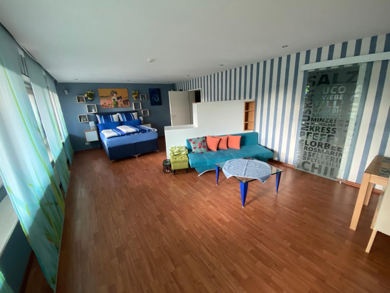 Ein modernes Wohnzimmer mit einem blauen Sofa, einem grünen Sessel und einem Glastisch. Ein Bett mit blauer Bettwäsche steht an der Wand, und es gibt ein großes Fenster mit Vorhängen.