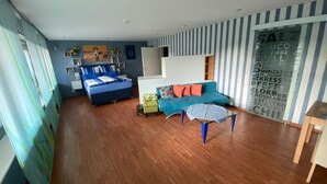 A modern living room with a blue sofa, a green armchair, and a glass coffee table. A bed with blue bedding is against the wall, and there is a large window with curtains. - Ilonas Home (Baden-Baden)