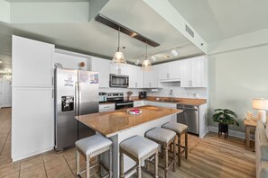 Una cocina moderna con armarios blancos, electrodomésticos de acero inoxidable, una isla de granito con taburetes altos, iluminación colgante y una pequeña planta en maceta.