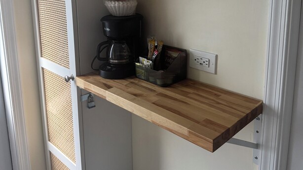 A kitchen with wooden shelves holding cups, a coffee maker, and a trash bin.