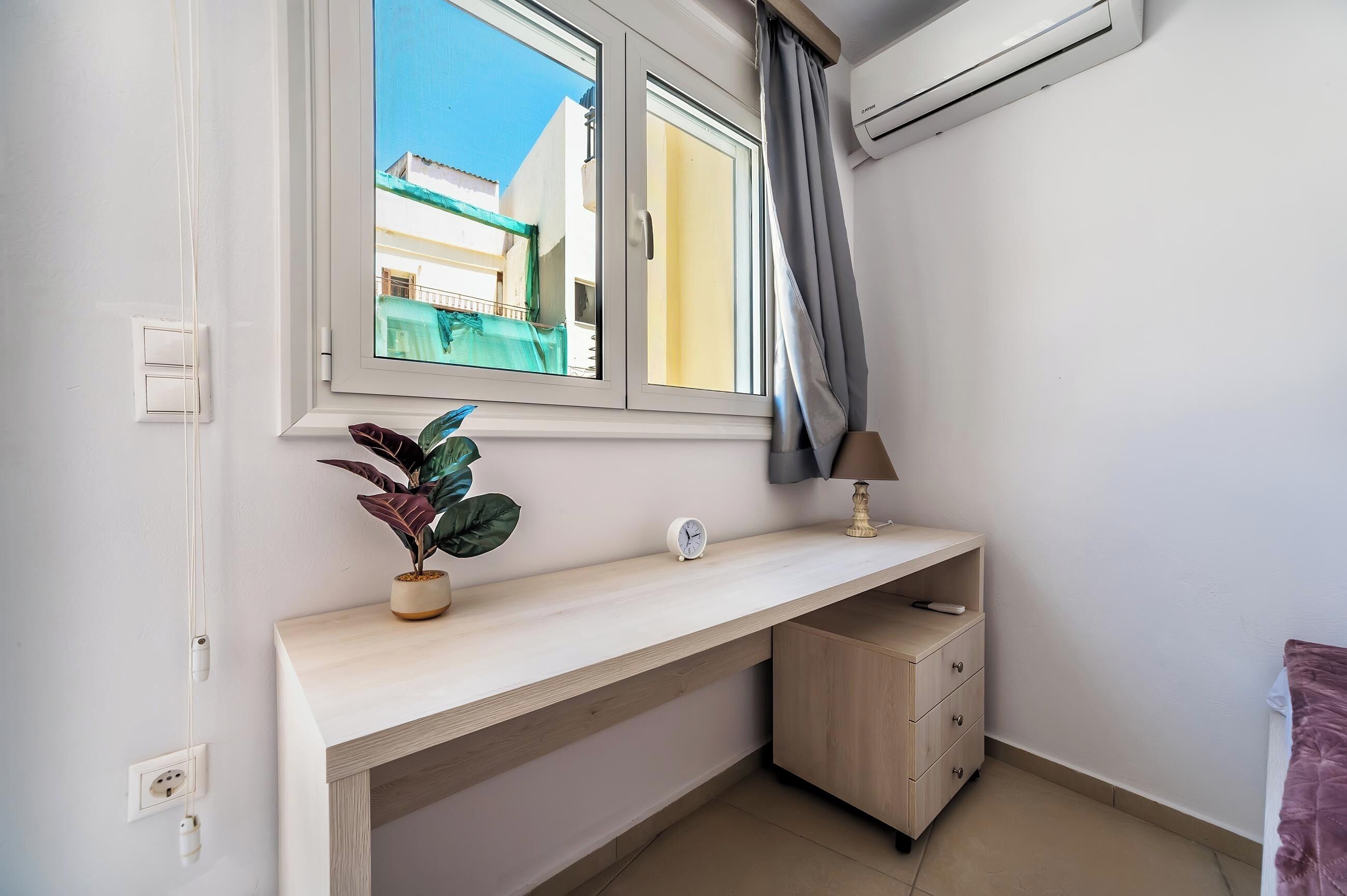 A bedroom with a window, a bedside table, a lamp, a plant, and a wardrobe.