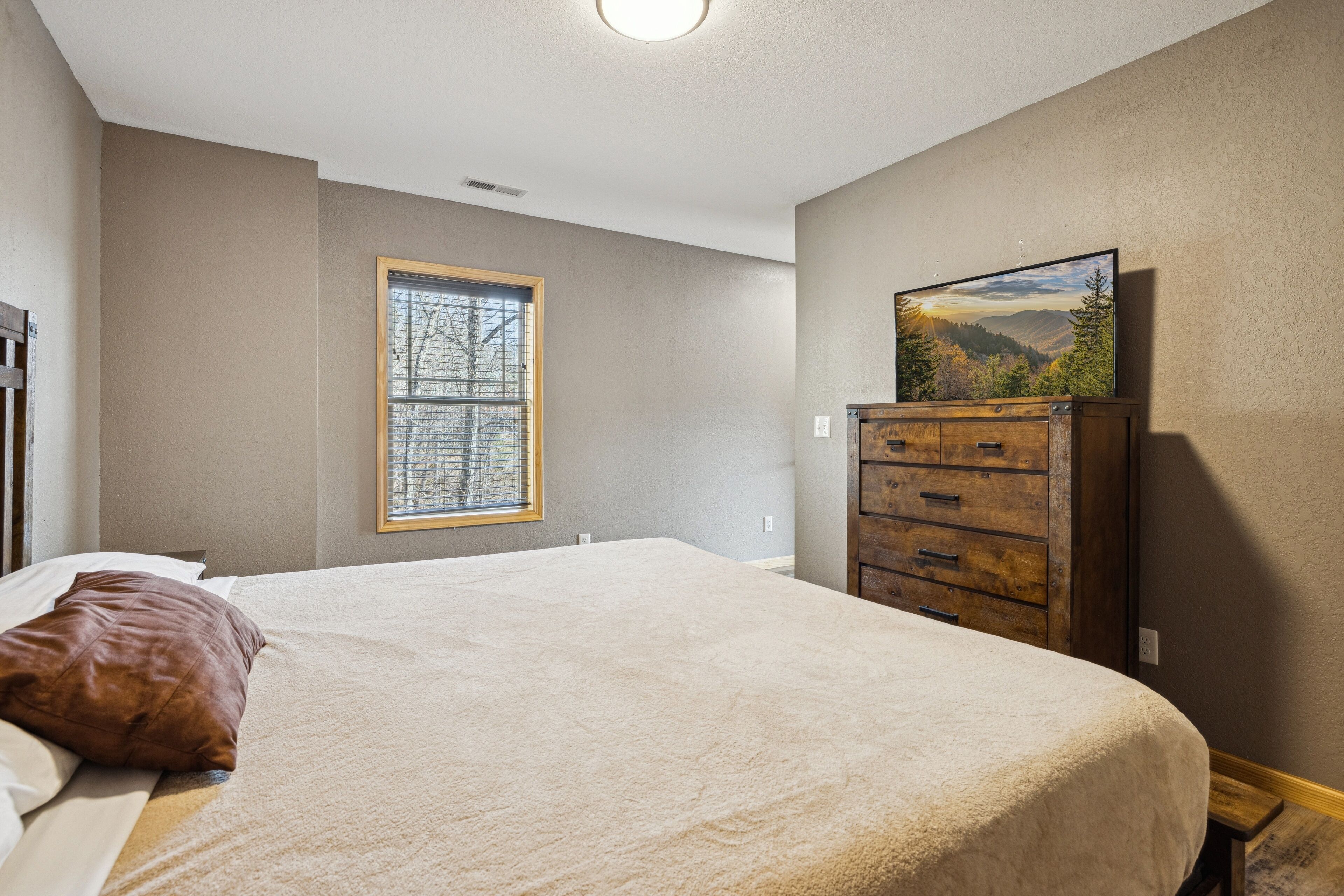A bedroom with a large bed, a wooden dresser, a window with blinds, and a painting on the wall.