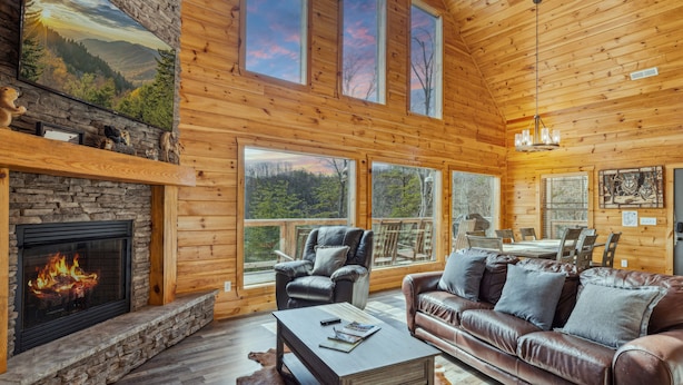 A cozy log cabin interior with a stone fireplace, leather furniture, and large windows offering scenic views. - Wild Heart Lodge 4 Bedroom Cabin (Sevierville)