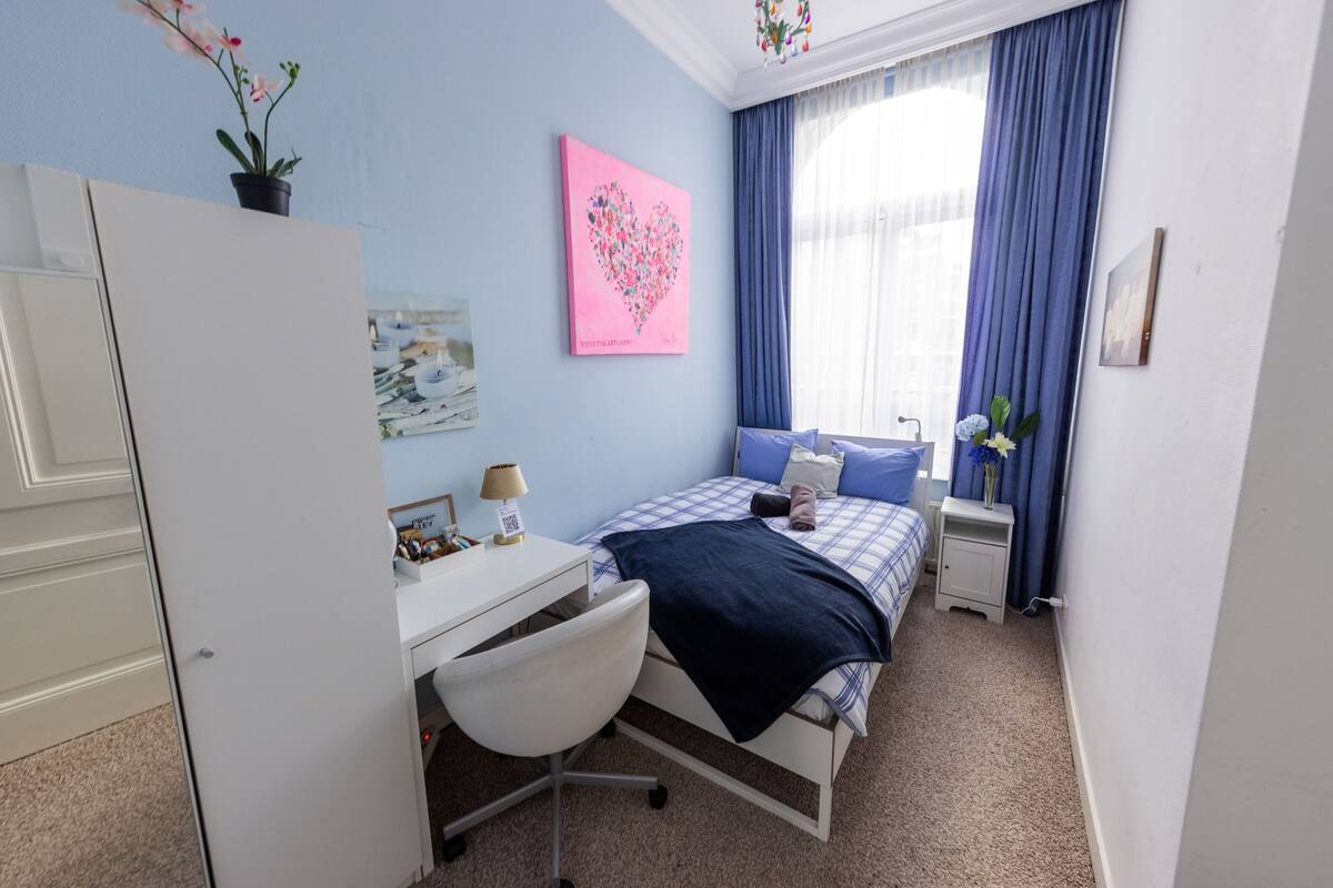 A bedroom with a bed, desk, chair, and a window with curtains.