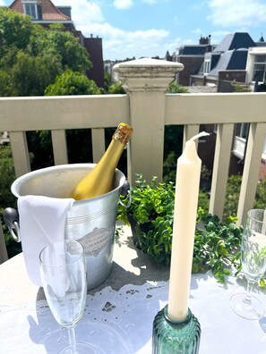 A table with a champagne bottle, a candle, and two glasses on a balcony with a railing and greenery in the background. - Flora's Palace  (The Hague)