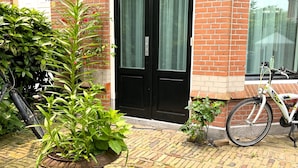 A brick building with a black door, a potted plant in the foreground, and a bicycle parked against the wall. - Flora's Palace (The Hague)