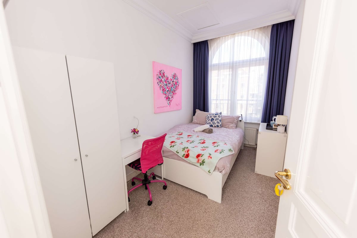 A bedroom with a bed, a desk, a chair, a window with curtains, and a wall with a heart-shaped artwork.