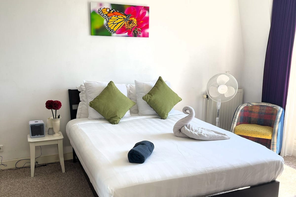 A neatly made bed with white bedding and green pillows, a wall-mounted fan, a small bedside table with a lamp, and a colorful butterfly painting above the bed.