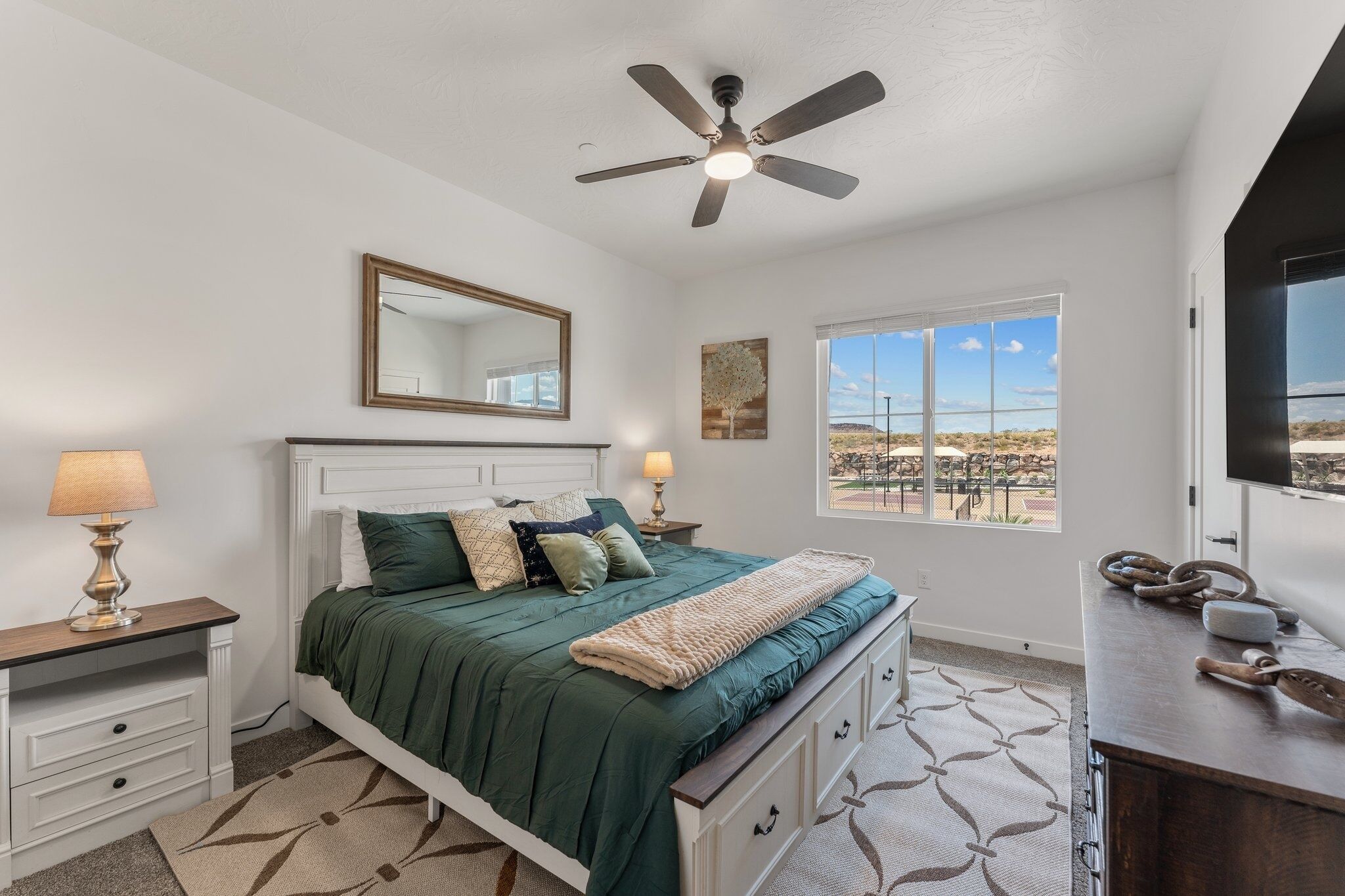 A bedroom with a bed, a ceiling fan, a large window with a view, a bedside table with a lamp, and a dresser.