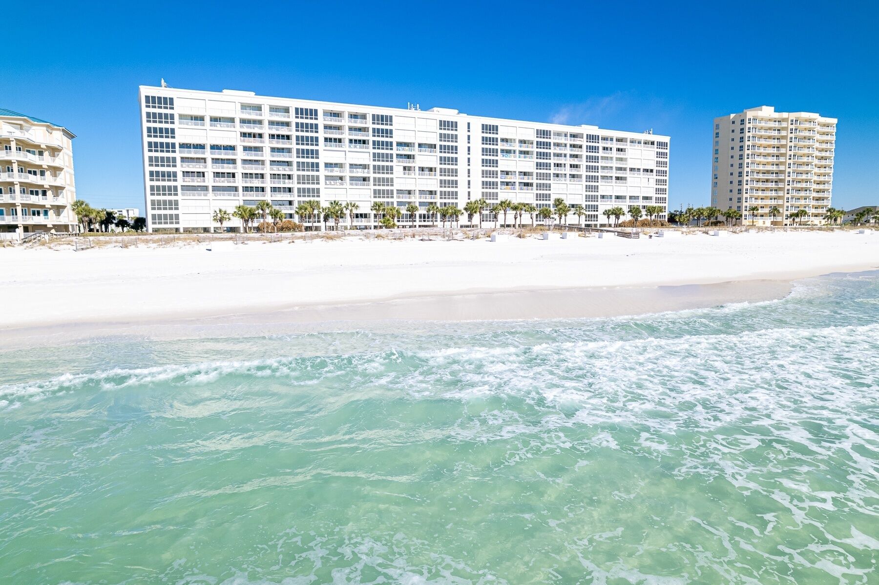 A beachfront hotel with multiple floors, a sandy beach, and palm trees.