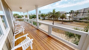 Une terrasse en bois, dotée de rampes et de chaises blanches, qui donne sur une rue bordée de palmiers et de bâtiments.