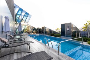 A modern outdoor swimming pool area with lounge chairs and umbrellas, surrounded by glass walls and contemporary architecture.