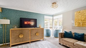 Una sala de estar con un televisor de pantalla plana grande que muestra 'NETFLIX', un mueble de madera, un sofá color beige y un ventanal con vistas al exterior.