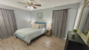 A bedroom with a bed, a ceiling fan, a nightstand with a lamp, a small potted plant, and a window with curtains.