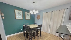 A dining area with a wooden table and chairs, teal walls, a clock, and a window with white curtains.