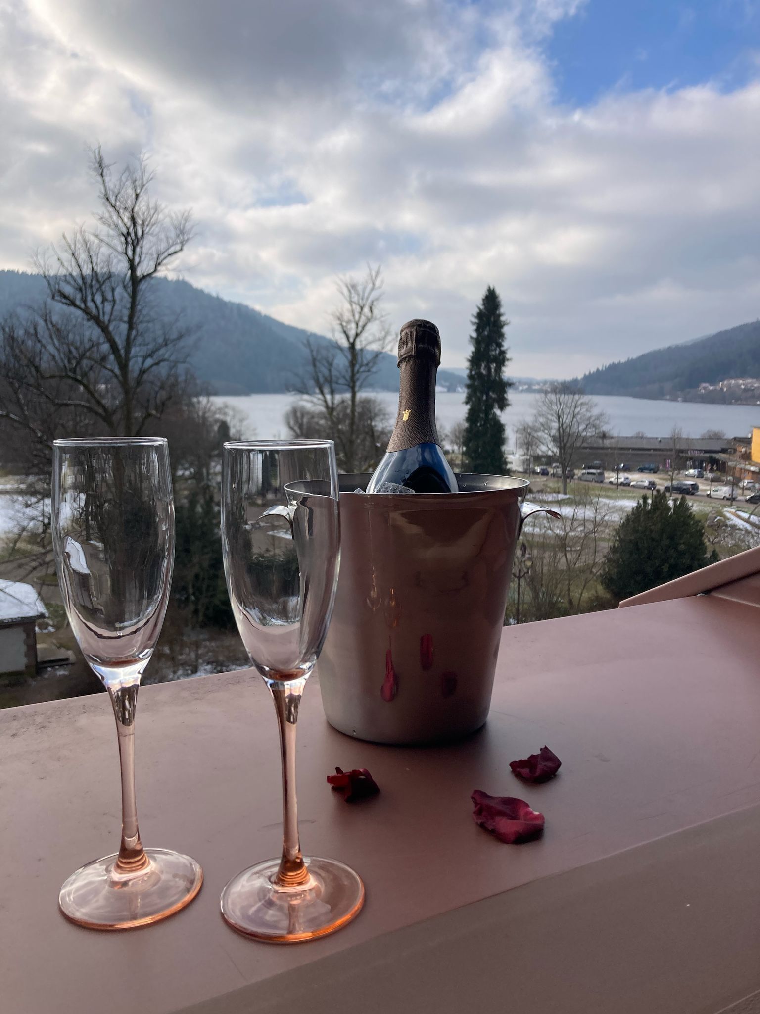 Two empty wine glasses on a table with a bottle of champagne in a bucket, set against a scenic view of a lake and mountains.